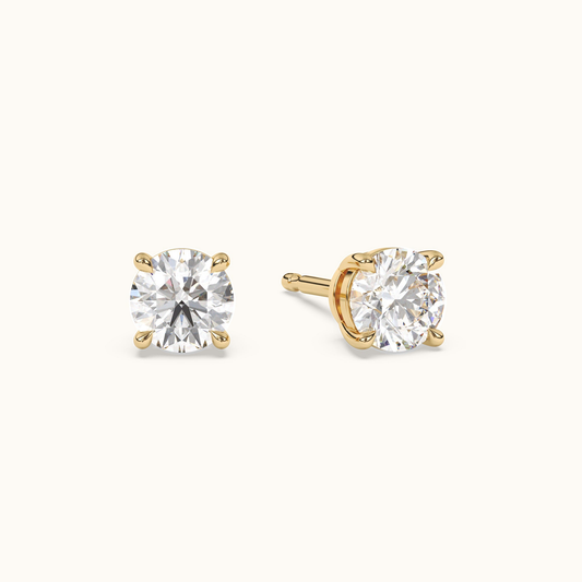 Round brilliant diamond studs in yellow gold, angled pair view on warm ivory
